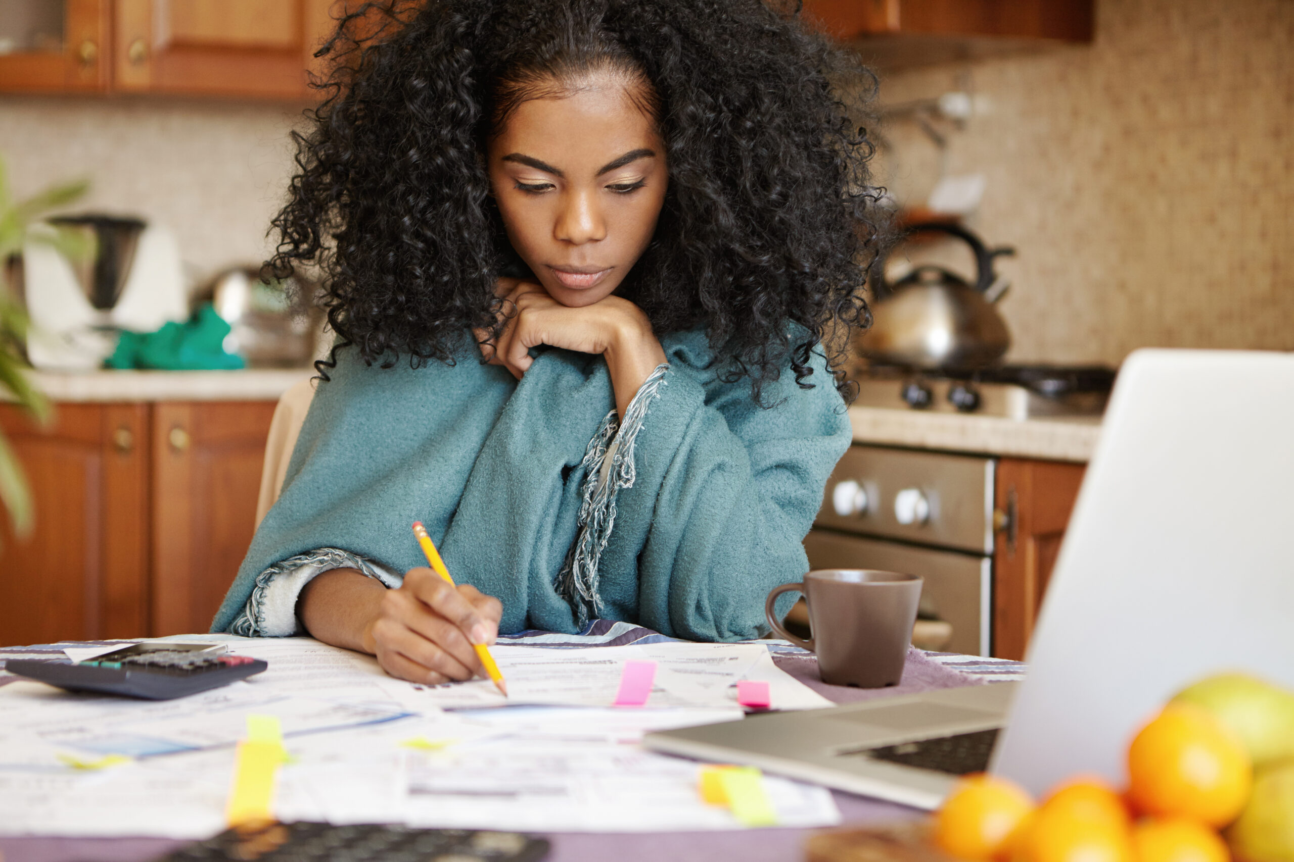 Unhappy depressed young dark-skinned housewife managing domestic budget alone at night, making notes with pencil, sitting at kitchen table with open laptop, documents, calculator and cup of coffee