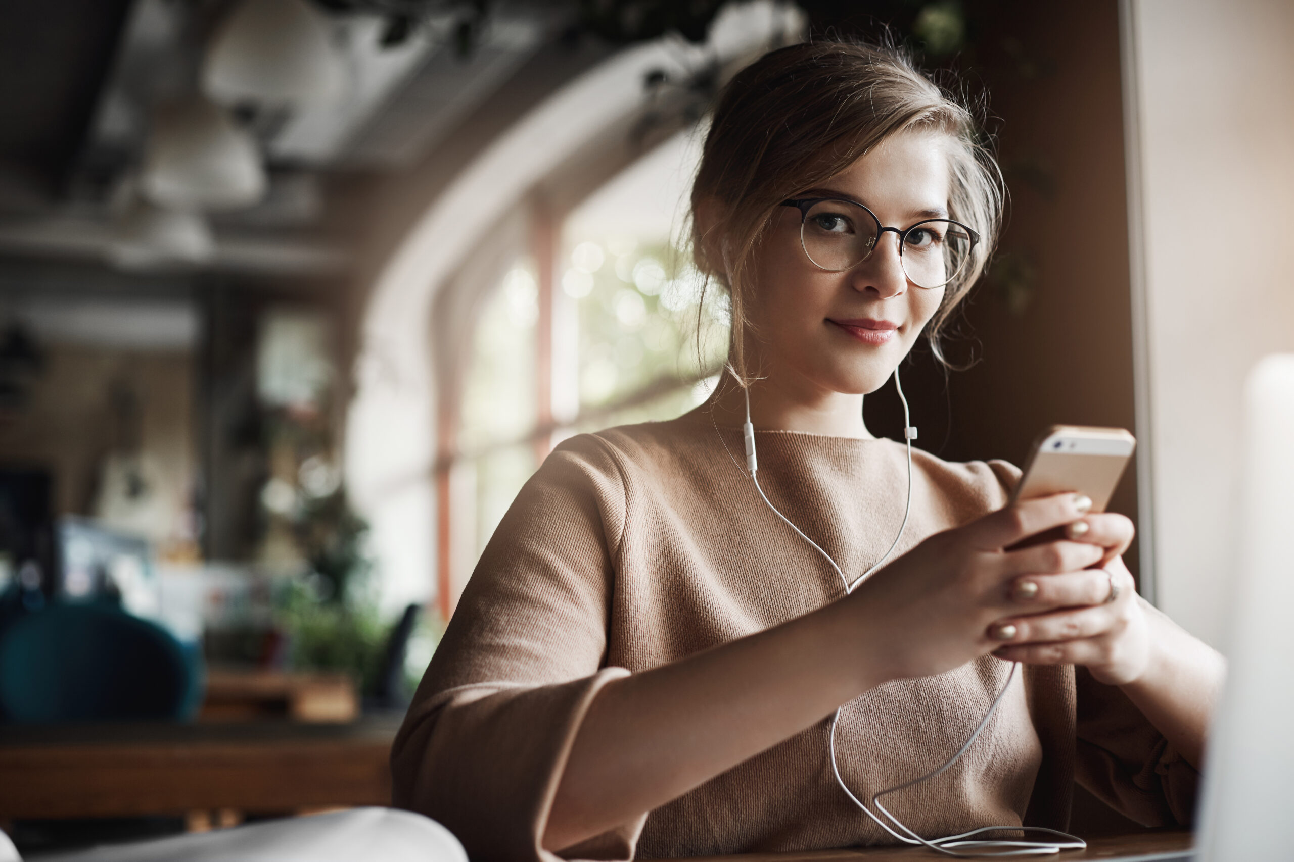 Indoor shot of good-looking stylish and happy caucasian female with fair hair in glasses, holding smartphone and wearing earbuds while watching video, distracting to look and smile at camera