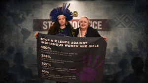 Jovem mulher indígena com cocar tradicional segura faixa denunciando a violência contra mulheres indígenas durante evento internacional.